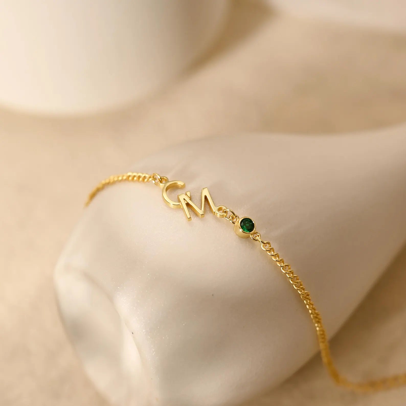 A delicate gold chain bracelet with a custom 'CM' initial pendant and a small green gemstone accent, displayed on a cream-colored jewelry stand.