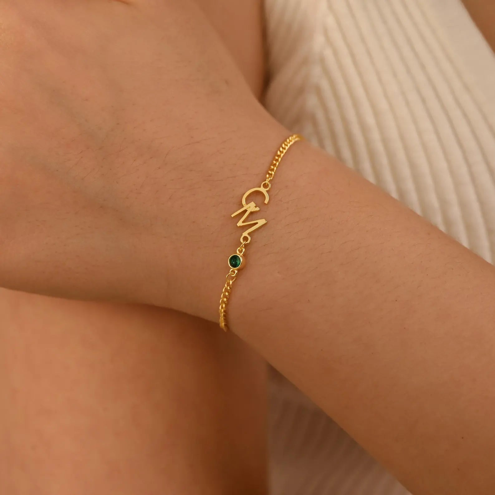 A delicate gold chain bracelet with a custom initial charm and a small green stone accent, shown worn on a person's wrist.