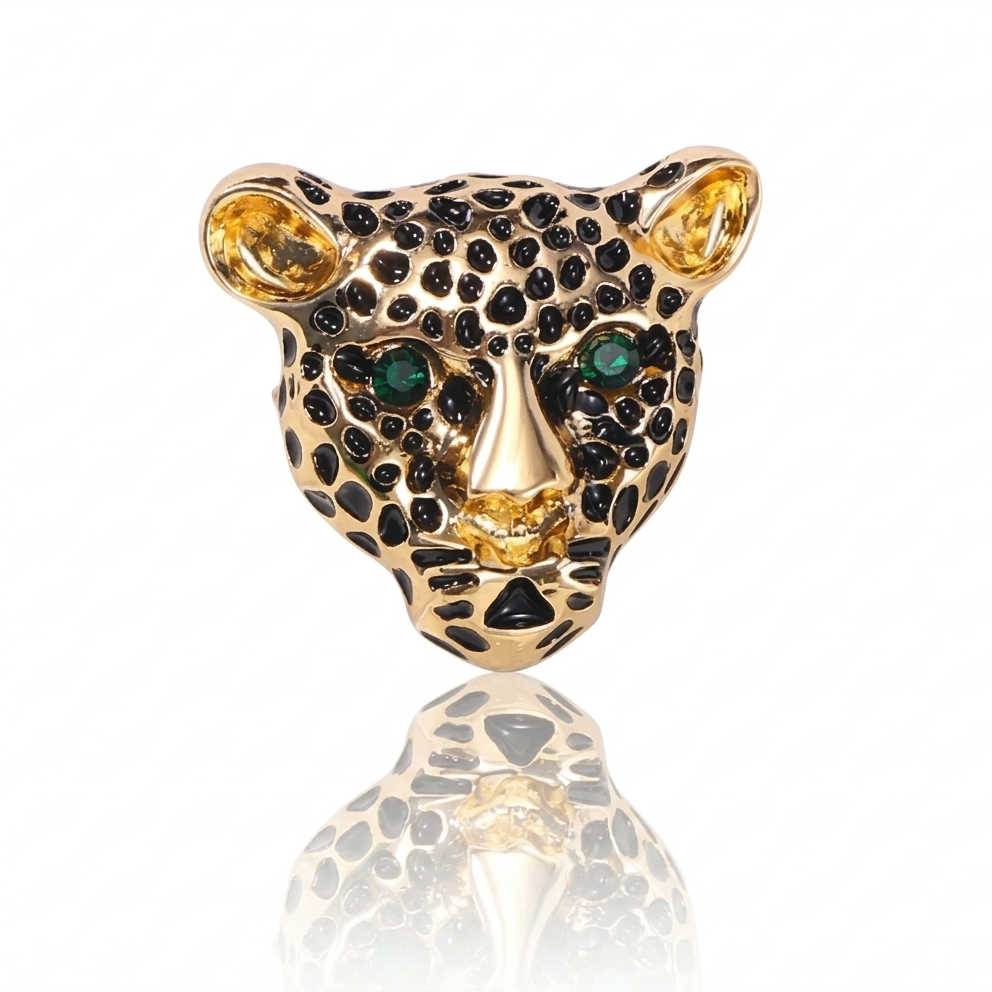 A leopard head charm or pendant in gold-tone metal, detailed with black enamel spots, green gemstone eyes, and a gold nose and ears, shown on a white background with a reflection below.