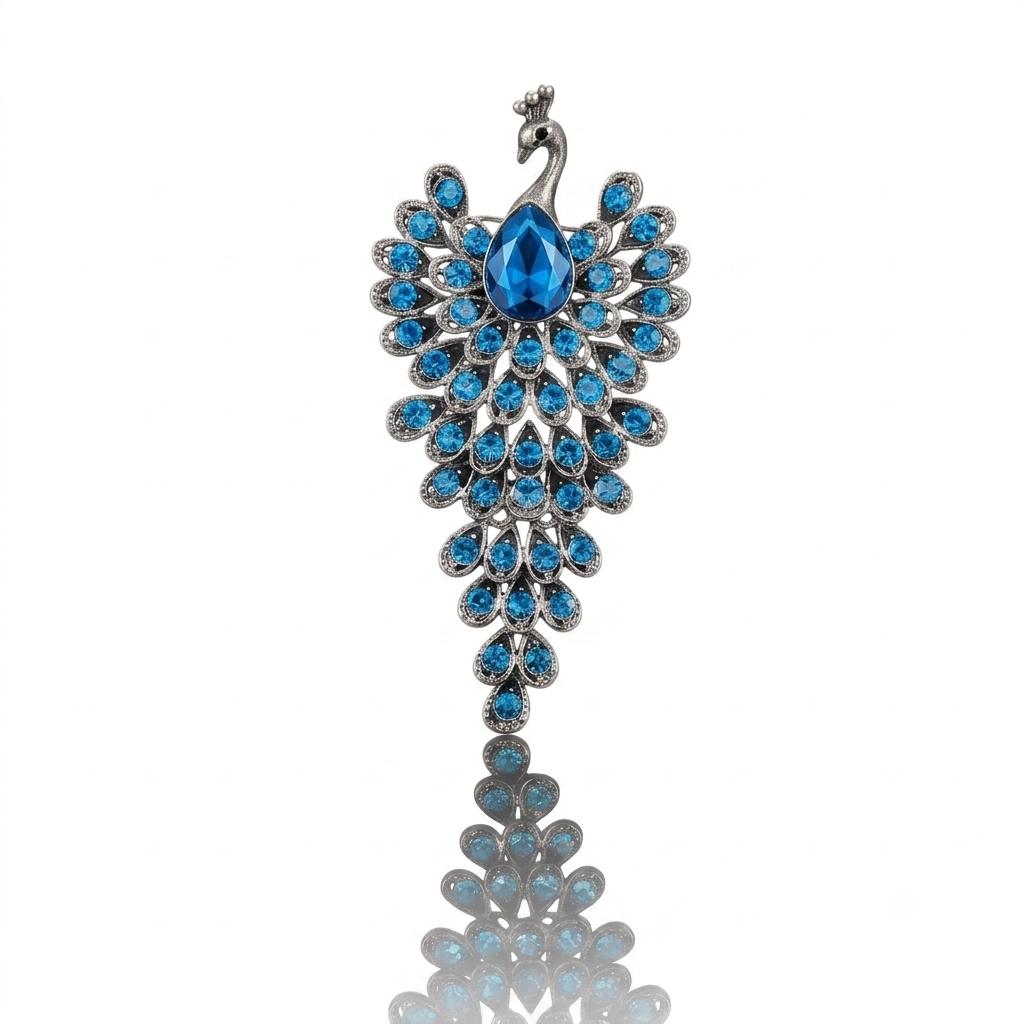 A peacock-themed brooch featuring a silver-toned metal setting adorned with vibrant blue teardrop and round rhinestones, with a central larger blue teardrop stone. The piece is shown against a plain white background with a soft reflection below.