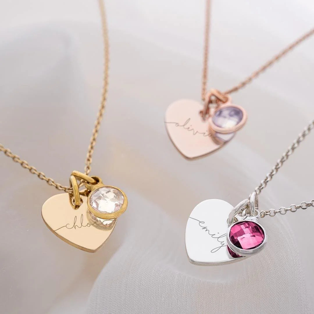 Three heart-shaped necklaces with engraved names and colored stones on a light background