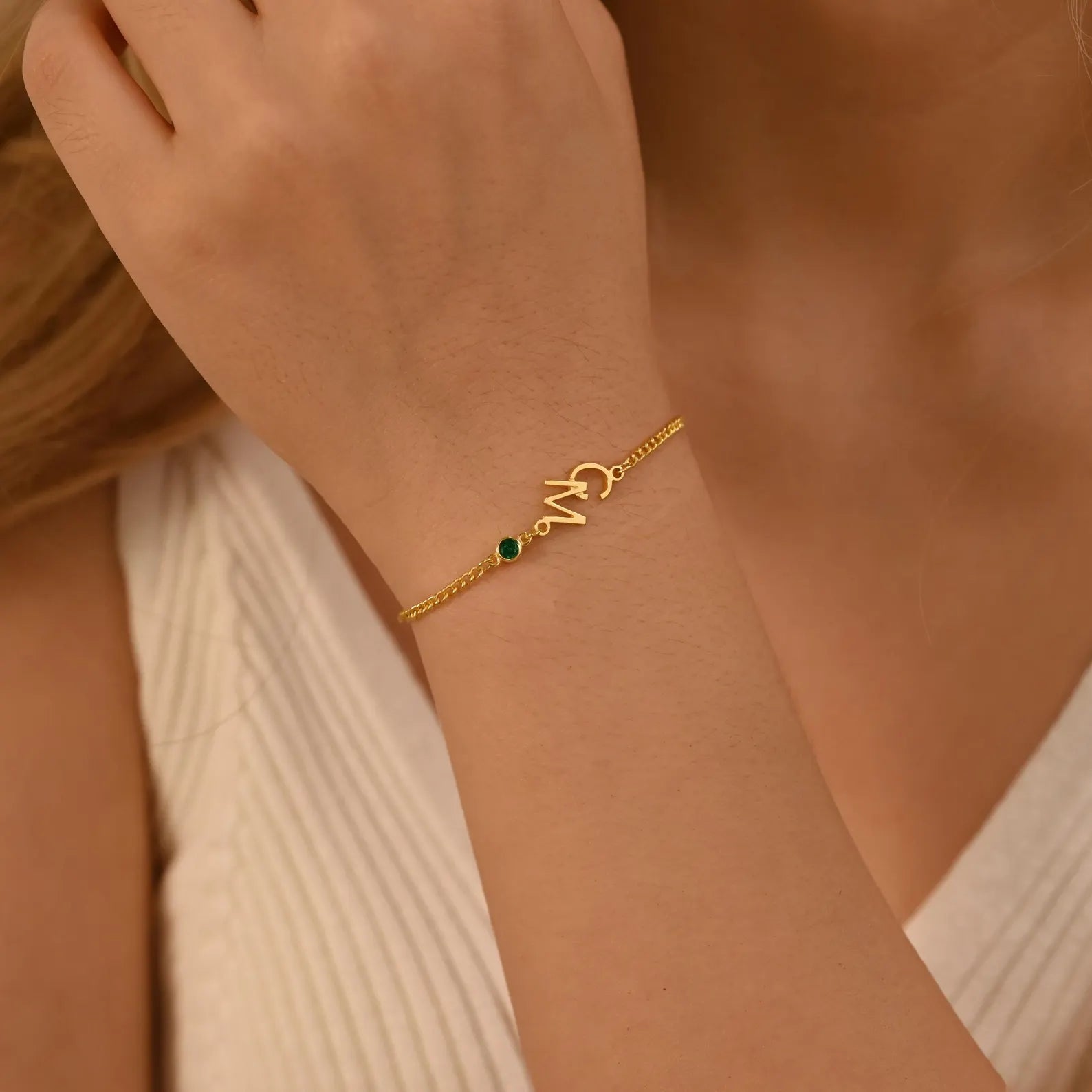 A close-up of a delicate gold chain bracelet worn on a woman's wrist. The bracelet features a small green oval stone and a gold initial charm spelling 'M'.