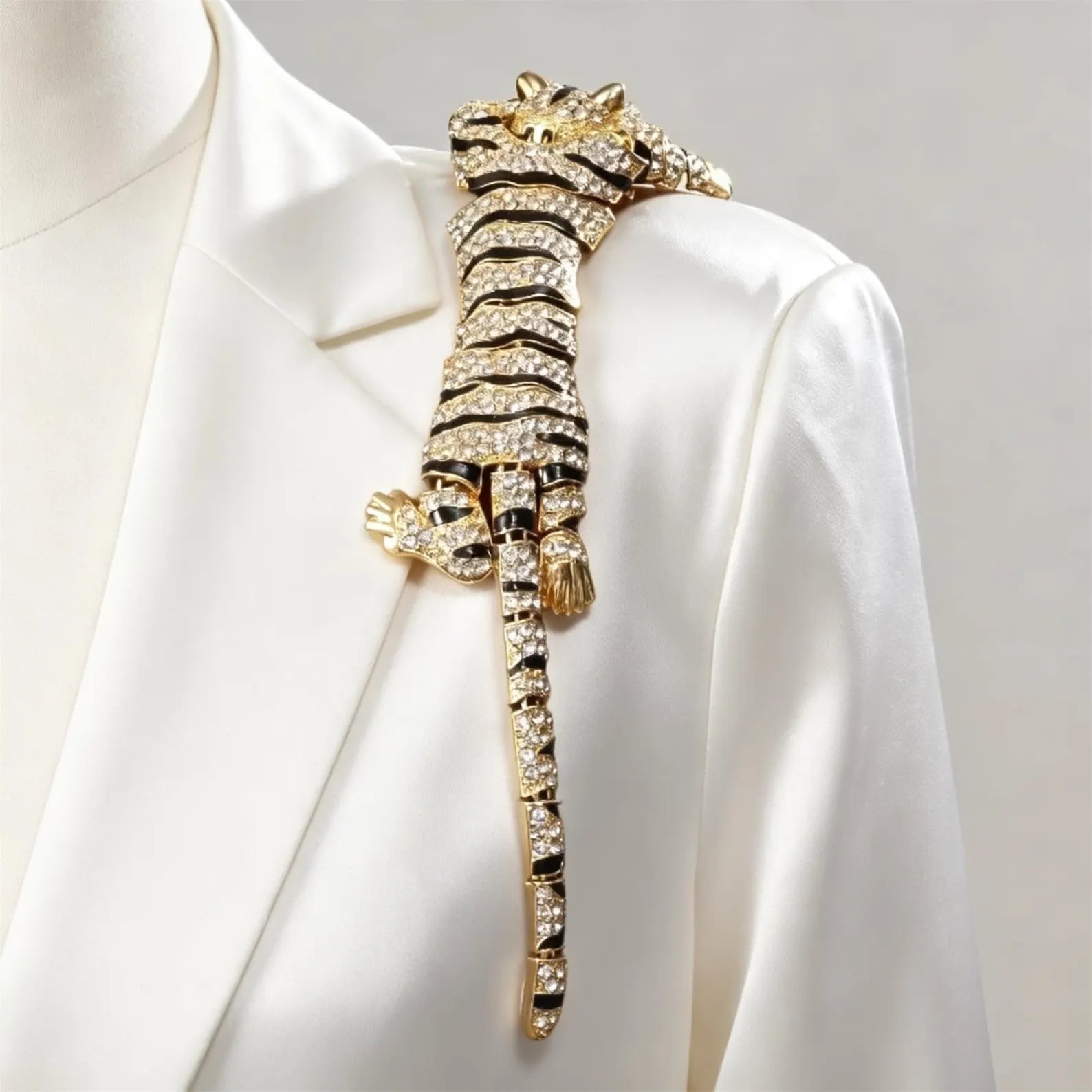 A gold-toned animal-shaped brooch with clear rhinestones, shaped like a tiger or cheetah, displayed on a white satin blazer against a light neutral background.