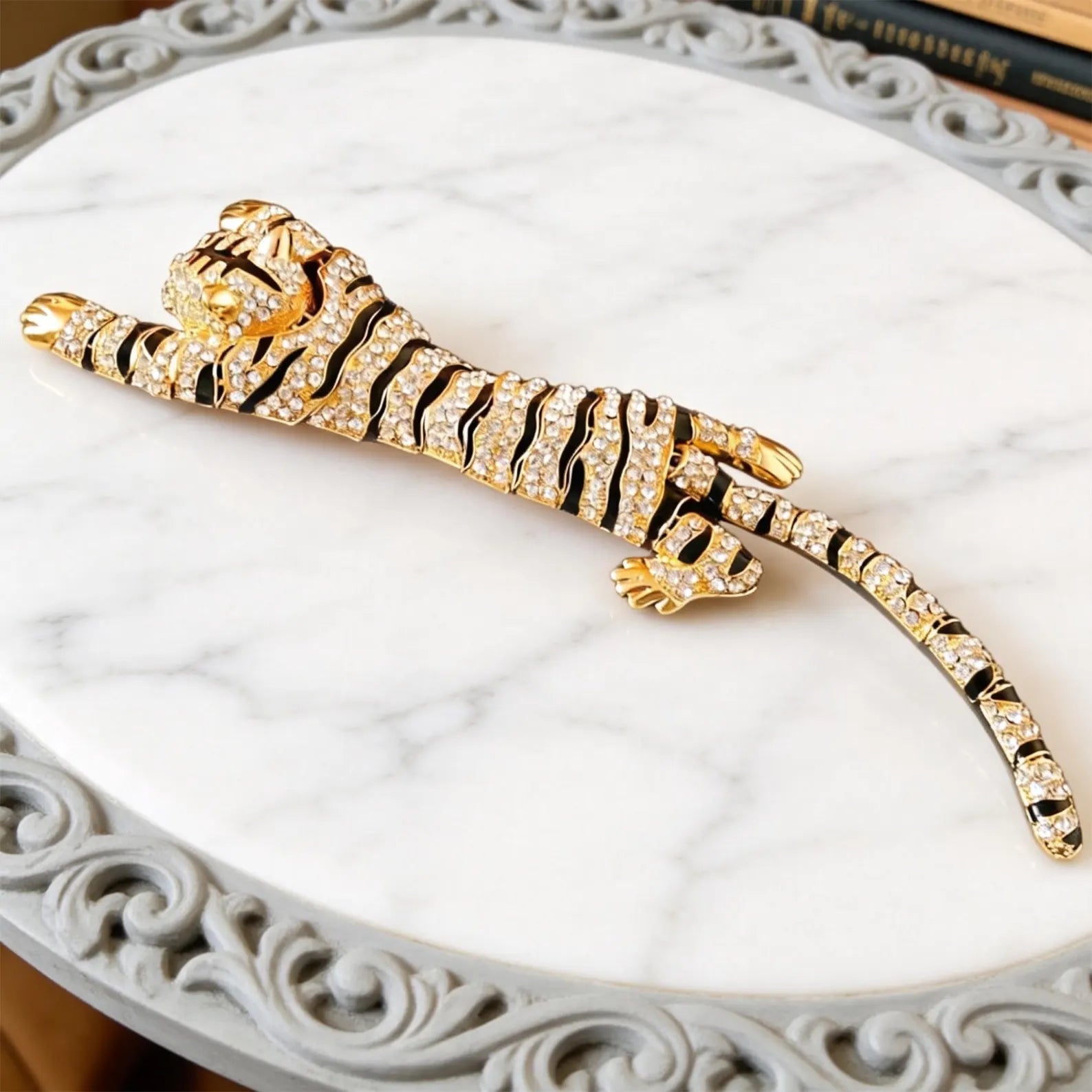 A gold tiger-shaped brooch or hair accessory covered with clear rhinestones, featuring black enamel stripes, displayed on a white marble round tray with an ornate gray border.