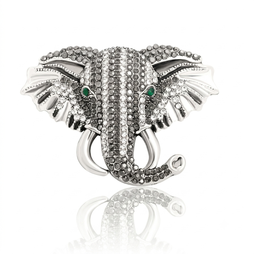 A silver-toned elephant head brooch or pin embellished with clear rhinestones and small green stone eyes, shown on a white background with a soft reflection below.