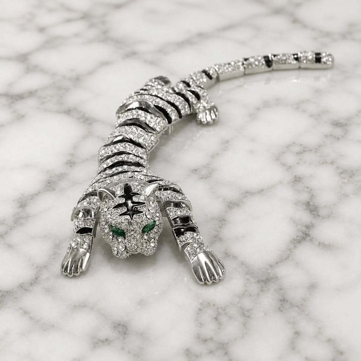 A silver tiger-shaped brooch or pendant covered in clear stones, with black enamel-style striping and green eye accents, displayed on a white marble surface.