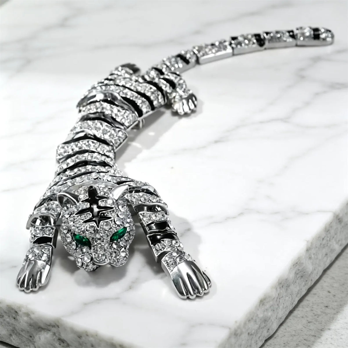 A silver tiger-shaped jewelry piece, heavily embellished with clear crystals and featuring green gemstone eyes, resting on a white marble surface.
