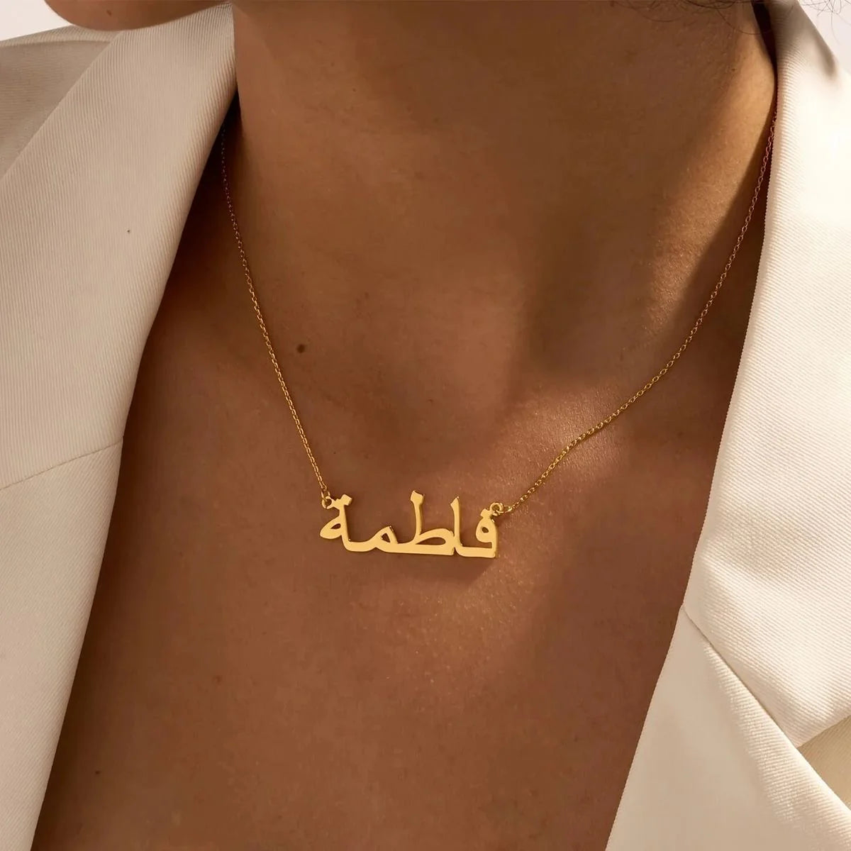 Gold necklace with Arabic script on a person wearing a beige blazer.