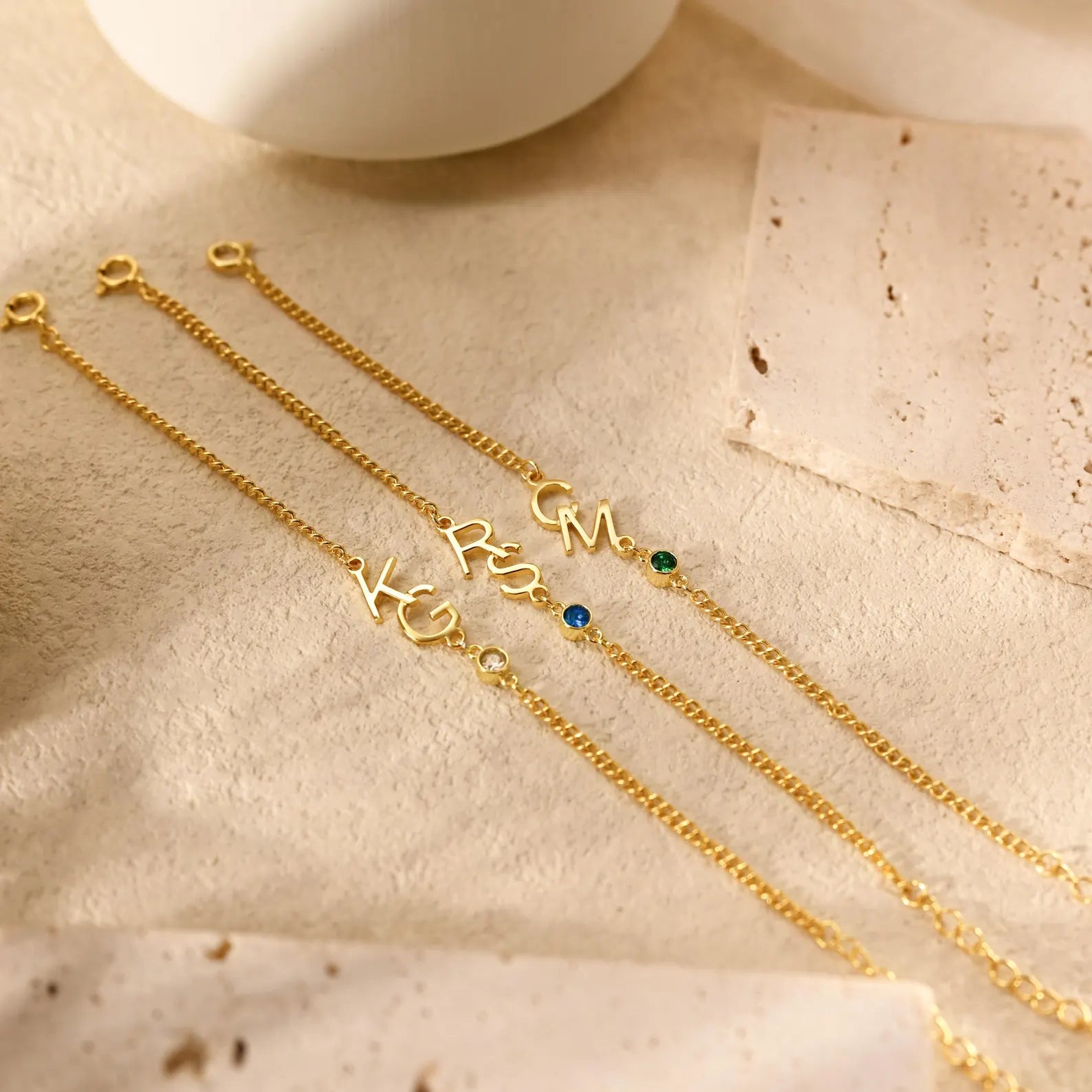 Three personalized gold chain necklaces with script initials and small gemstone accents, arranged on a textured beige surface.