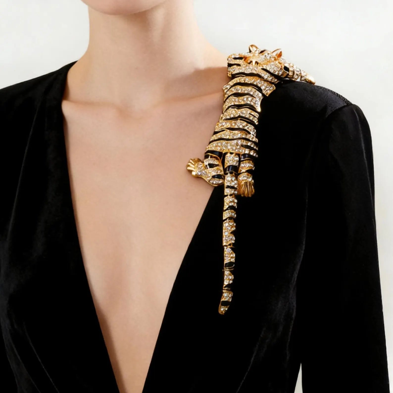 A close-up of a model wearing a black velvet deep V-neck dress, featuring a large, ornate gold crocodile brooch embellished with clear crystals on the shoulder.