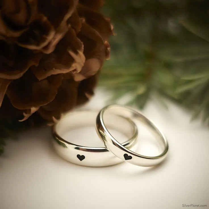 Two silver rings with heart designs on a white surface with a pine cone and branch in the background.