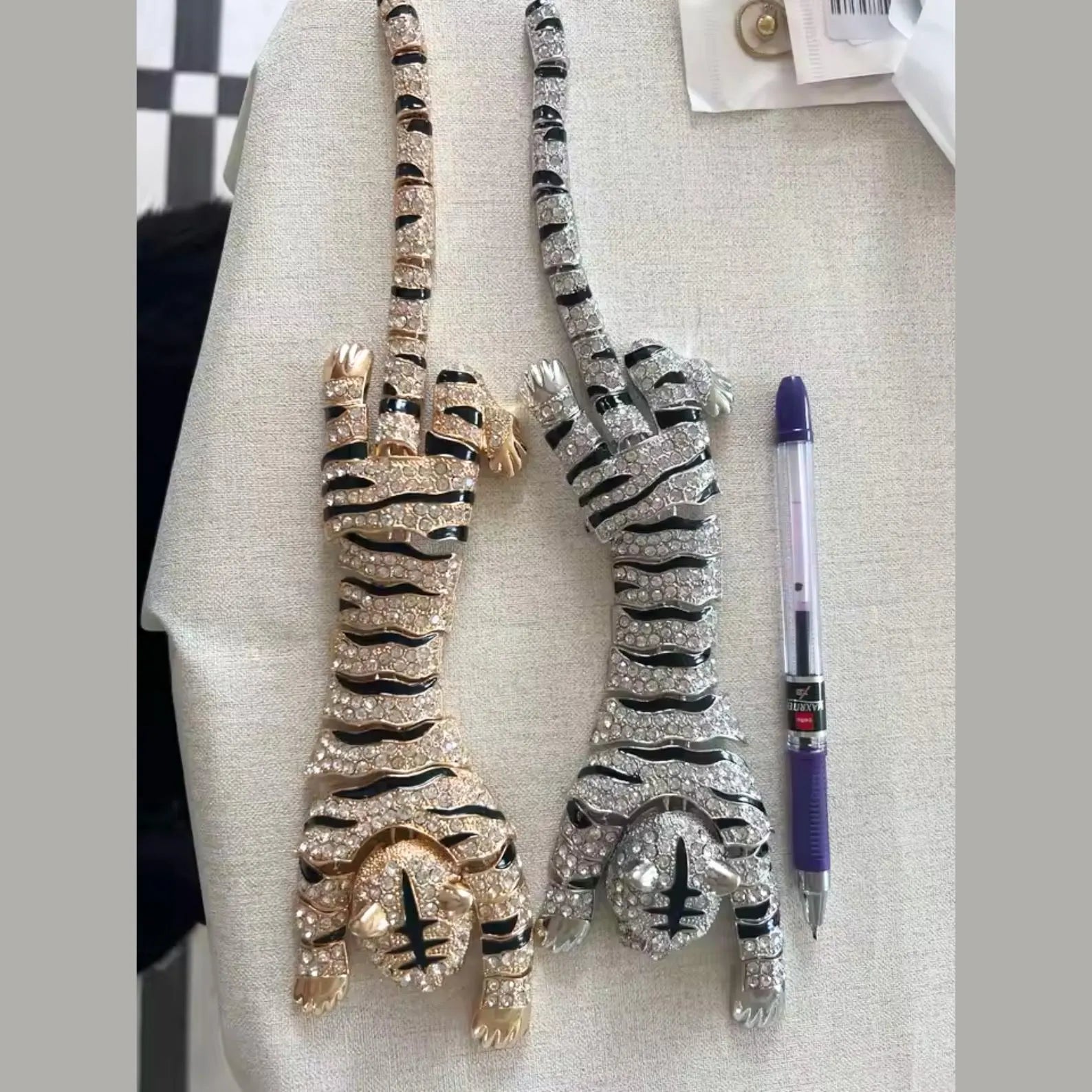 Two tiger-shaped handbag charms or keychains covered in rhinestones, one in gold and one in silver, displayed on a beige fabric surface next to a purple pen for scale.