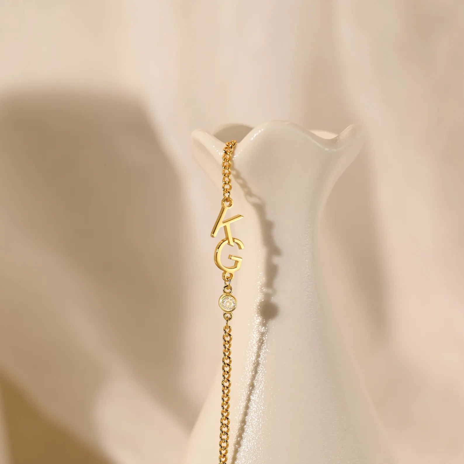 A delicate gold chain necklace with a personalized initial pendant reading 'KAG' and a small round charm, displayed on a white ceramic vase against a soft beige fabric background.